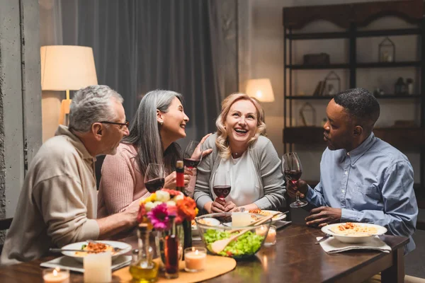 Smiling multicultural friends talking and holding wine glasses during dinner — Stock Photo