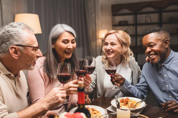 Smiling multicultural friends talking and clinking with wine glasses during dinner — Stock Photo