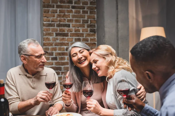 Amigos multiculturais sorridentes abraçando e segurando copos de vinho durante o jantar — Fotografia de Stock