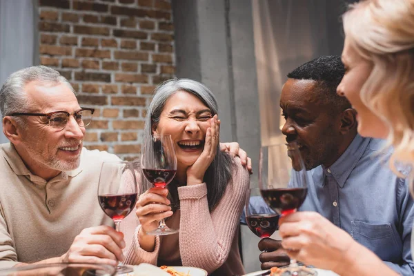 Smiling multicultural friends hugging and holding wine glasses during dinner — Stock Photo