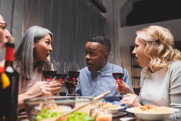 Shocked multicultural friends talking and holding wine glasses during dinner — Stock Photo
