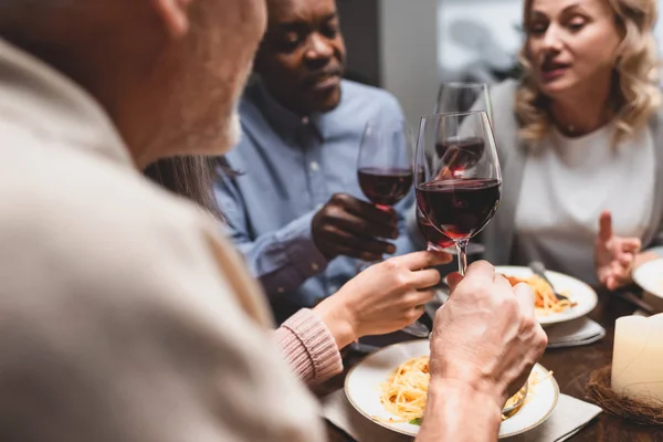 Vista recortada de amigos multiculturales hablando y sosteniendo copas de vino durante la cena - foto de stock