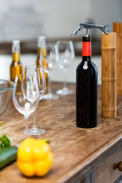 Enfoque selectivo de verduras, sacacorchos, botellas con vino y cerveza, copas de vino, molino de pimienta, molino de sal en la mesa de madera en la cocina - foto de stock