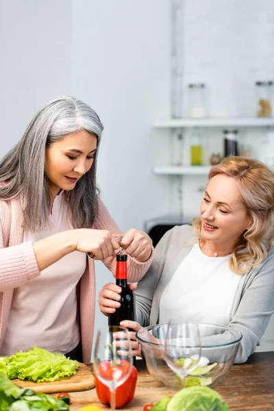 Souriant amis multiculturels ouverture bouteille de vin avec tire-bouchon dans la cuisine — Photo de stock