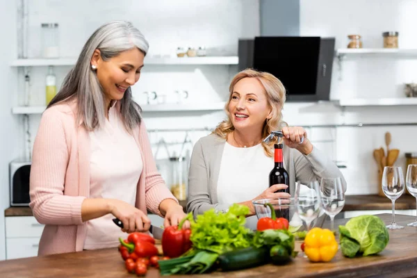 Smiling woman opening wine bottle with corkscrew and looking at her asian friend with knife — Stock Photo