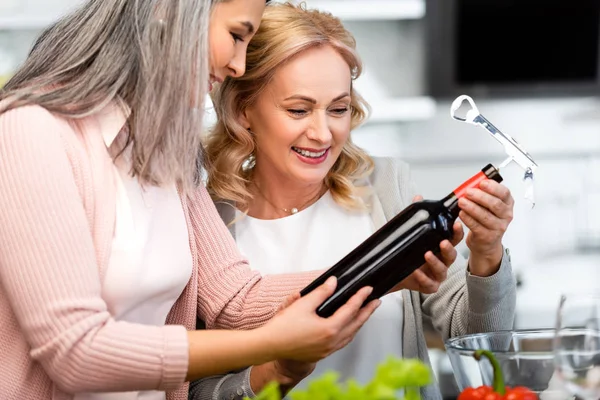 Smiling multicultural friends looking at wine bottle with corkscrew in kitchen — Stock Photo