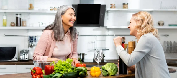 Plano panorámico de mujer sonriente abriendo botella de vino con sacacorchos y mirando a su amigo asiático - foto de stock