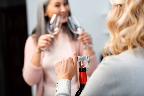 Vista recortada de la mujer abriendo botella de vino con sacacorchos y su amigo asiático sosteniendo copas de vino - foto de stock