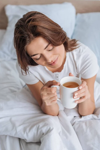 Vista aérea de chica feliz con los ojos cerrados sosteniendo taza con té en la cama - foto de stock