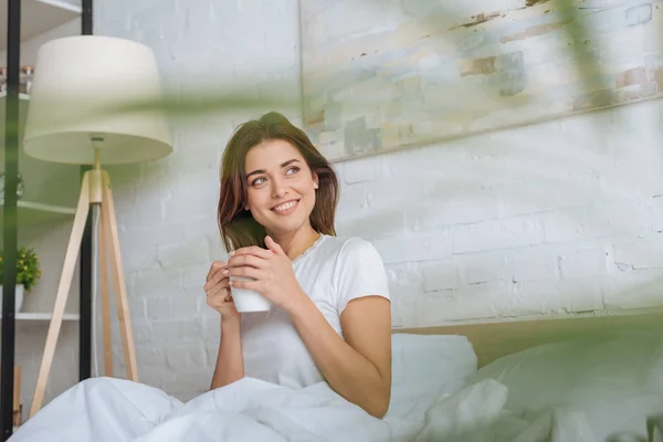 Messa a fuoco selettiva di felice giovane donna guardando lontano mentre tiene in mano la tazza con il tè a letto — Foto stock