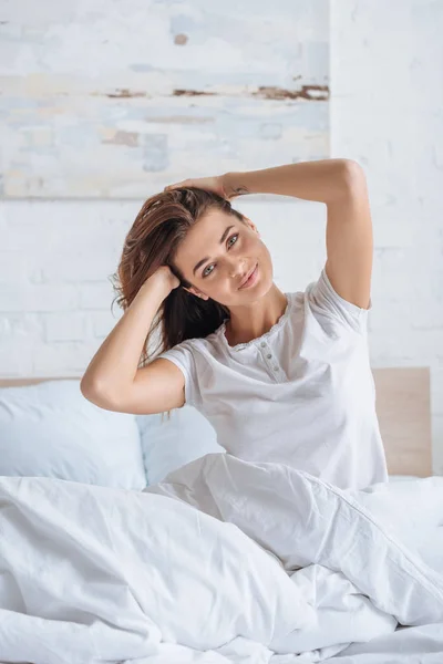 Donna felice toccare i capelli e guardando la fotocamera a letto — Foto stock