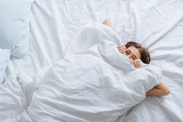 Top view of young woman covering face with blanket while resting in bed — Stock Photo