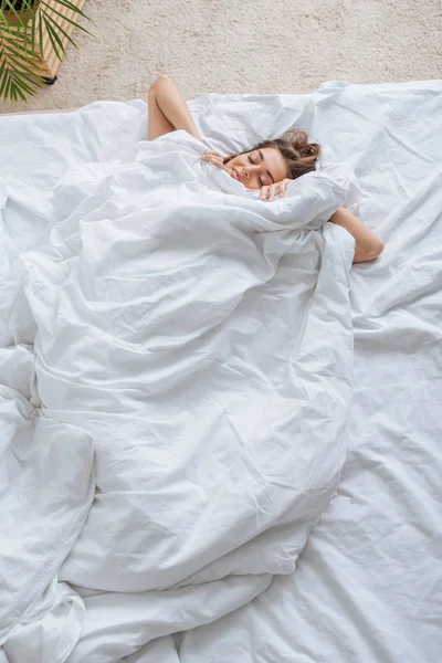 Вид сверху на счастливую молодую женщину, покрытую одеялом, отдыхающую в постели — стоковое фото