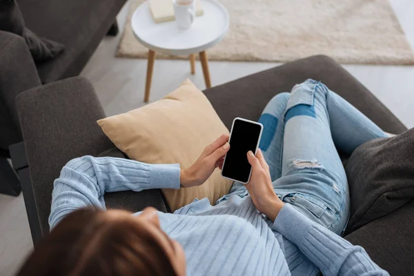 Vista aérea de la mujer sosteniendo el teléfono inteligente con pantalla en blanco mientras se enfría en casa - foto de stock