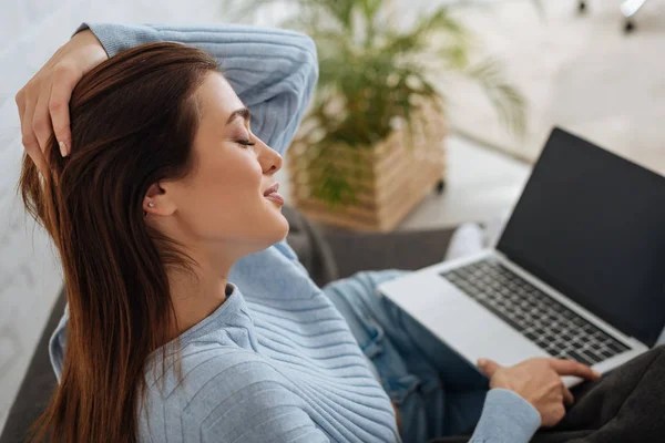 Atractiva chica con los ojos cerrados sosteniendo portátil con pantalla en blanco en casa - foto de stock