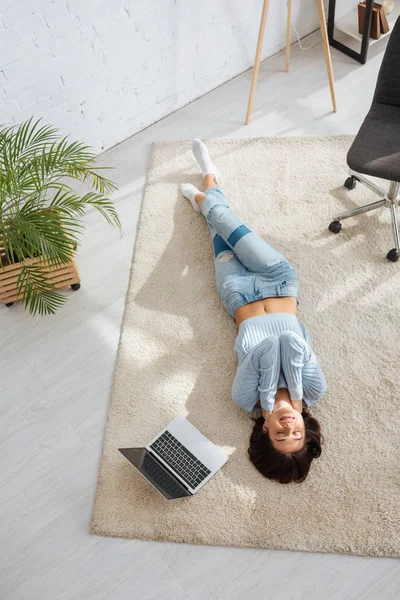 Vista superior de la chica alegre que se enfría mientras está acostado en la alfombra cerca de la computadora portátil en casa - foto de stock