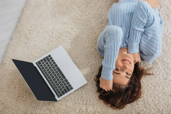 Vista superior da menina feliz refrigeração enquanto deitado no tapete perto do laptop em casa — Fotografia de Stock