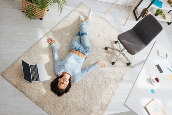 Vista superior da mulher positiva descansando enquanto deitado no tapete perto do laptop em casa — Fotografia de Stock