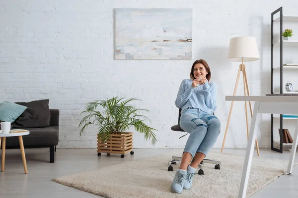 Fröhliches Mädchen, das Tasse hält und zu Hause auf Stuhl sitzt — Stockfoto