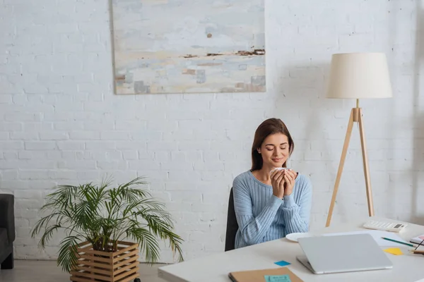 Junge verträumte Frau hält Tasse in der Hand und lächelt am heimischen Tisch — Stockfoto