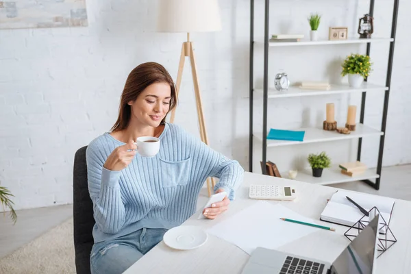 Femme heureuse en utilisant un smartphone tout en tenant une tasse de café et de refroidissement à la maison — Photo de stock