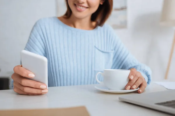 Vista recortada de la mujer alegre usando teléfono inteligente cerca de taza con café - foto de stock