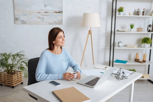 Glückliche Frau mit Tasse Kaffee in der Nähe von Gadgets auf dem Tisch — Stockfoto