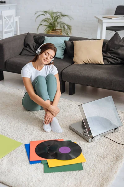 Jovem mulher ouvindo música em fones de ouvido perto de sofá e vintage gravador — Fotografia de Stock