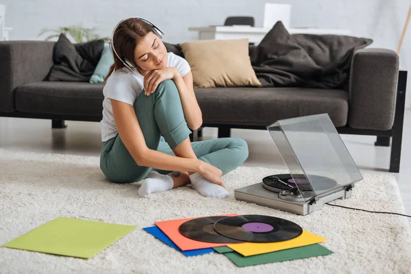 Dreamy young woman listening music in headphones near sofa and vintage record player — Stock Photo