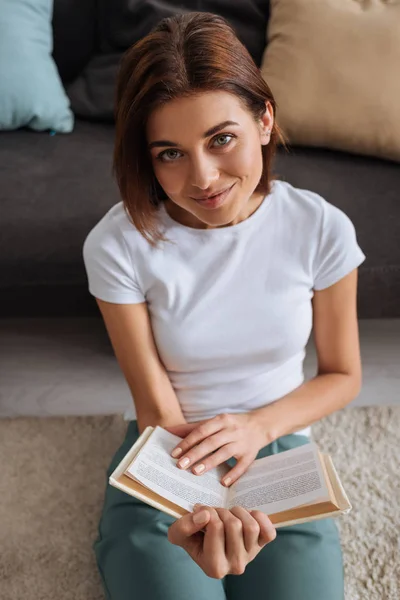 Happy woman holding book while sitting on carpet near sofa — Stock Photo