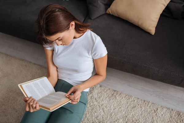 Vista aerea di ragazza lettura libro mentre seduto sul tappeto vicino divano — Foto stock