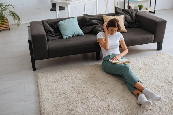 Cheerful young woman reading book while chilling near sofa in living room — Stock Photo