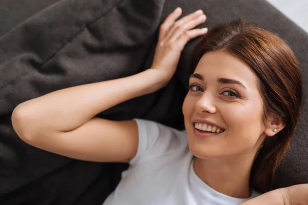 Draufsicht einer glücklichen Frau, die in die Kamera schaut, während sie auf dem Sofa im Wohnzimmer ruht — Stockfoto