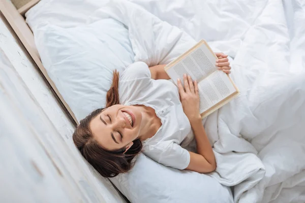 Vue de dessus de fille heureuse tenant livre tout en refroidissant au lit — Photo de stock