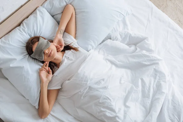 Vista superior da jovem mulher em máscara ocular descansando na cama em casa — Fotografia de Stock