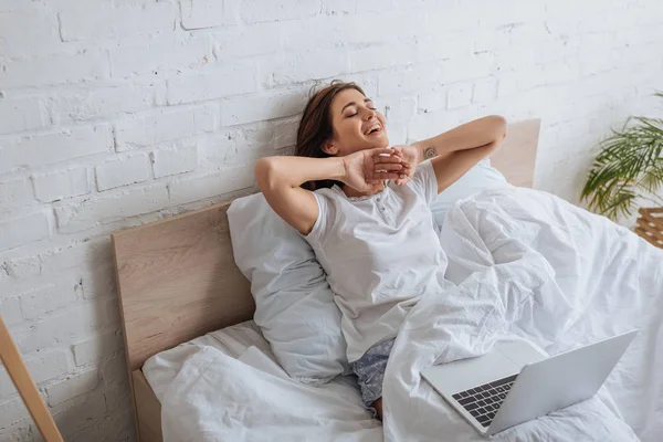 Dreamy woman smiling while chilling in bed near laptop — Stock Photo