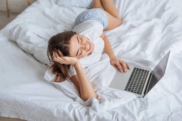 Mujer alegre usando el ordenador portátil mientras se enfría en el dormitorio - foto de stock