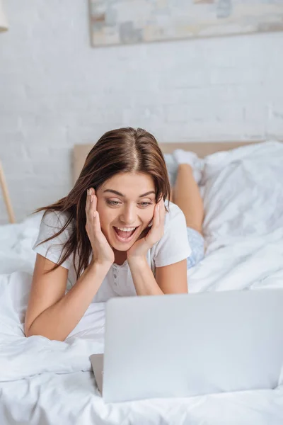 Возбужденная молодая женщина с помощью ноутбука во время охлаждения в спальне — стоковое фото