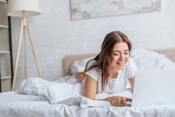 Mujer joven feliz usando el ordenador portátil mientras se enfría en el dormitorio - foto de stock
