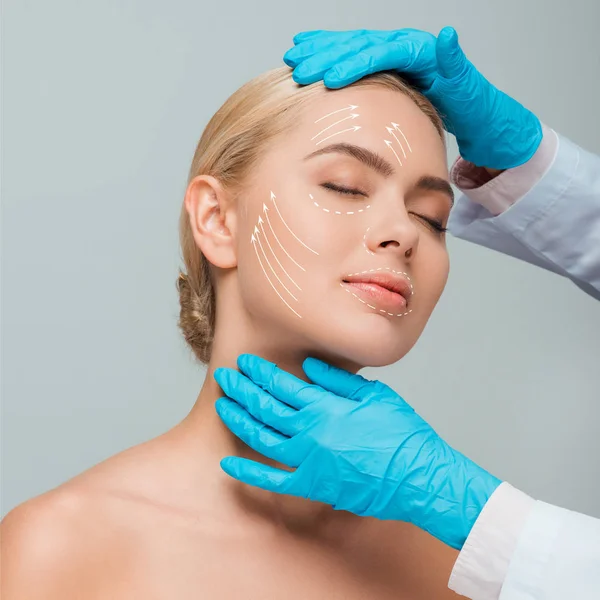 Vista recortada de esteticista en guantes de látex azul tocando mujer desnuda con ojos cerrados y marcas de cirugía plástica aisladas en gris - foto de stock