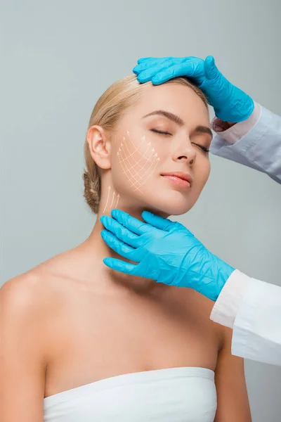 Vista recortada de esteticista en guantes de látex azul tocando a mujer con los ojos cerrados y marcas de cirugía plástica en la cara aislada en gris - foto de stock