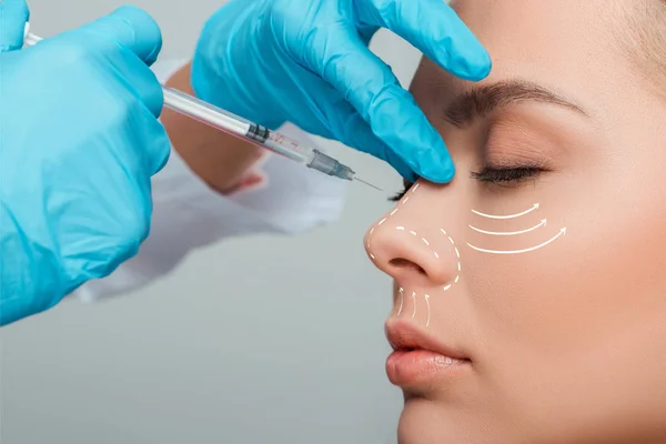 Vista recortada del cosmetólogo haciendo inyección de belleza en la nariz a una mujer atractiva aislada en gris - foto de stock