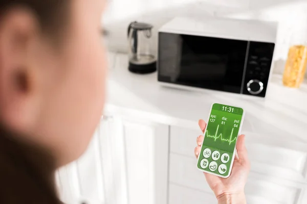 Selective focus of woman holding smartphone with health app and microwave on background — Stock Photo