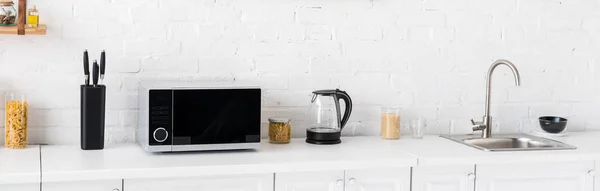 Panoramic shot of microwave, electric kettle, pasta, knives and sink in kitchen — Stock Photo