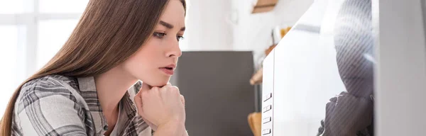 Panoramic shot of pensive woman looking at microwave in kitchen — Stock Photo