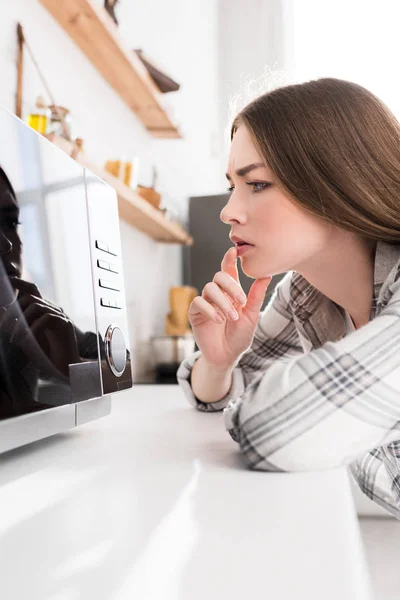 Pensive woman in shirt looking at microwave in kitchen — Stock Photo
