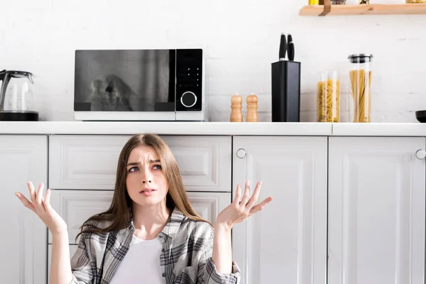 Mujer confundida haciendo gesto de encogimiento cerca de microondas - foto de stock