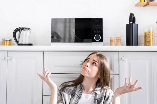 Mujer confundida y atractiva mirando al microondas y haciendo gesto encogiéndose de hombros en la cocina - foto de stock