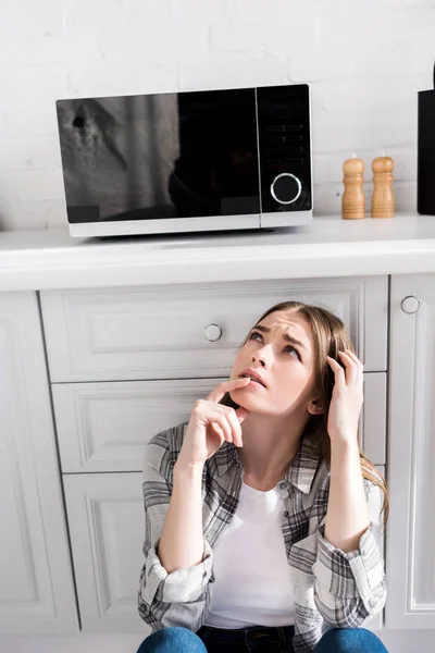 Mujer pensativa y atractiva mirando microondas en la cocina - foto de stock