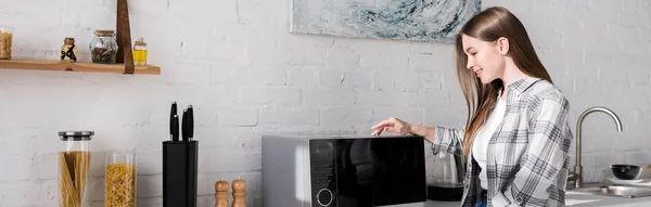 Panoramic shot of smiling woman looking at microwave in kitchen — Stock Photo